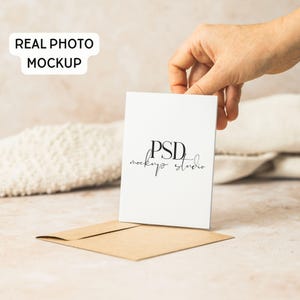 May include: A white card with the text "PSD mockup studio" is held above a brown envelope. The words "REAL PHOTO MOCKUP" are in a white rectangle in the upper left corner. The background is a neutral beige.