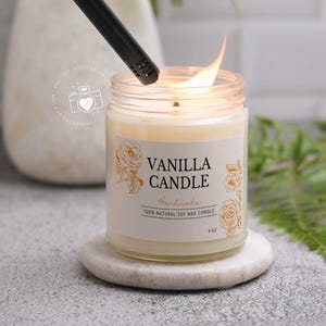 Op de afbeelding: Een witte kaars met een gouden label waarop "Vanilla Candle Handmade 100% Natural Soy Wax Candle 9 oz" staat, wordt aangestoken met een aansteker. De kaars staat op een witte marmeren onderzetter.