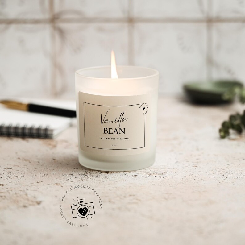 May include: A lit vanilla bean soy wax blend candle in a frosted glass jar. The label reads "Vanilla Bean" with a heart design. The candle is 3 inches tall and 3 inches wide. The background includes a notebook, pen, and a small green dish.