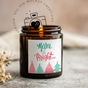 May include: A brown glass jar candle with a lit wick. The label reads "Merry & Bright" in green and pink script, with illustrated Christmas trees. The candle is on a textured surface.