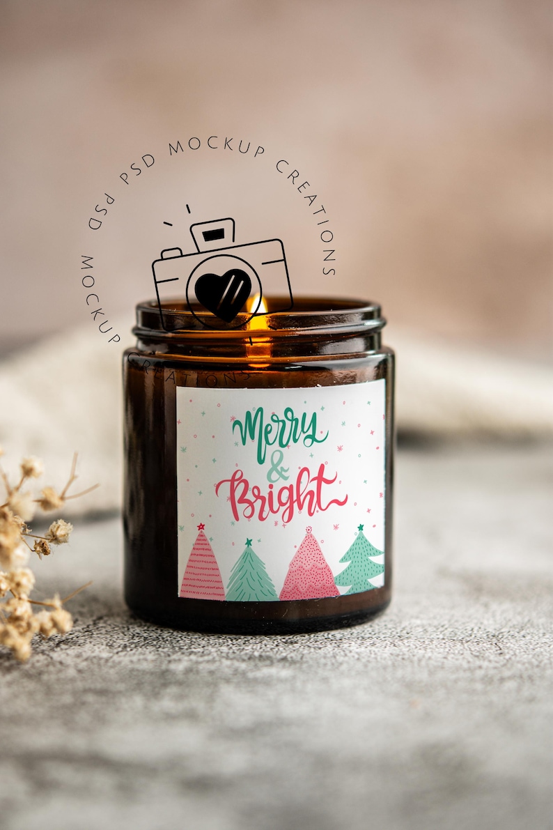 May include: A brown glass jar candle with a lit wick. The label reads "Merry & Bright" in green and pink script, with illustrated Christmas trees. The candle is on a textured surface.