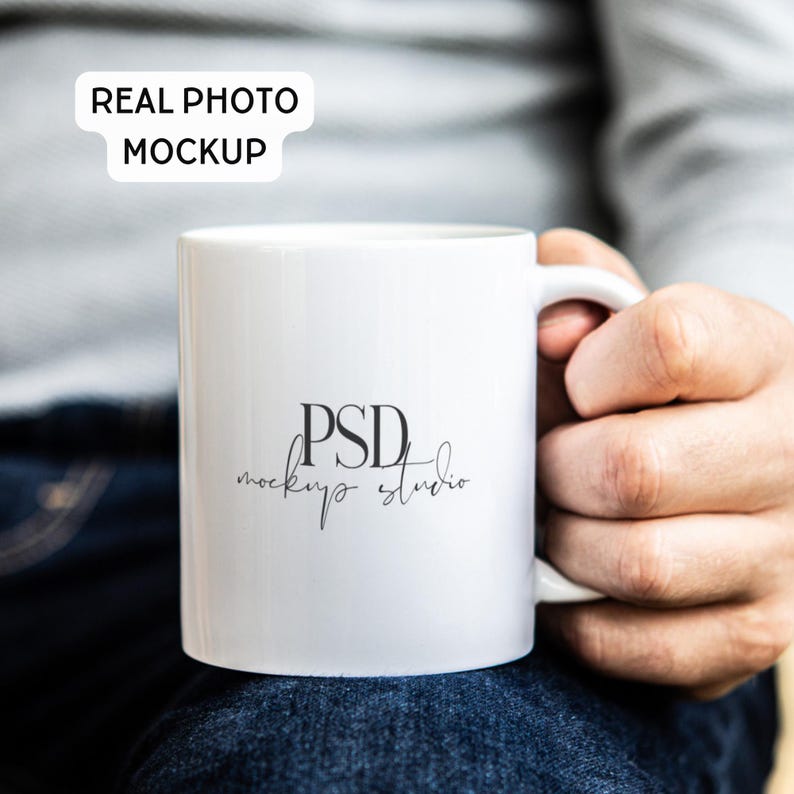 May include: White ceramic mug with a handle, featuring the text "PSD mockup studio" in black. The mug is held by a person wearing a gray shirt and blue jeans. A white rectangle with the text "REAL PHOTO MOCKUP" is in the top left corner.