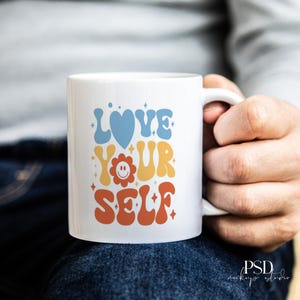 May include: White ceramic mug with the words "LOVE YOUR SELF" in a retro font. The word "LOVE" is in blue, "YOUR" is in yellow, and "SELF" is in orange. A blue heart and a smiling flower are also on the mug.