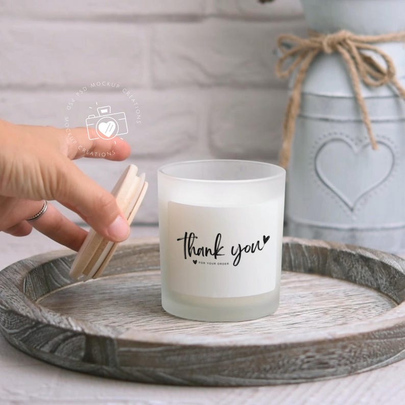May include: A white frosted glass candle jar with a white label that says "Thank you" with a small heart. The candle is on a wooden tray.
