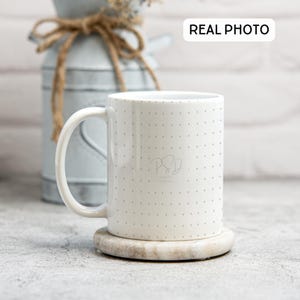 Puede incluir: Taza de cerámica blanca con un pequeño patrón de lunares grises. La taza tiene un logotipo negro y gris que dice "PSD" con el texto "Paper & Design" debajo. La taza está sentada sobre un posavasos redondo de piedra gris claro.