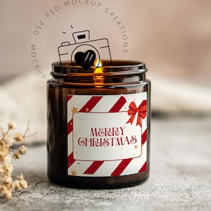 May include: A brown glass jar candle with a label that reads "MERRY CHRISTMAS" in red script. The label has red and white diagonal stripes and a red bow. The candle is lit, and the background is blurred.