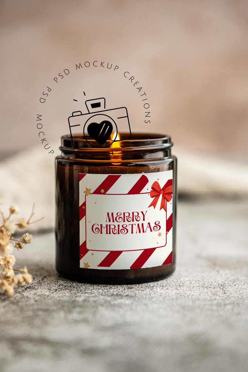 May include: A brown glass jar candle with a label that reads "MERRY CHRISTMAS" in red script. The label has red and white diagonal stripes and a red bow. The candle is lit, and the background is blurred.
