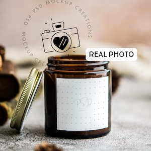 May include: A brown glass candle jar with a white label featuring a small logo and a dotted pattern. The lid is off and sitting next to the jar. The jar is on a light gray textured surface.