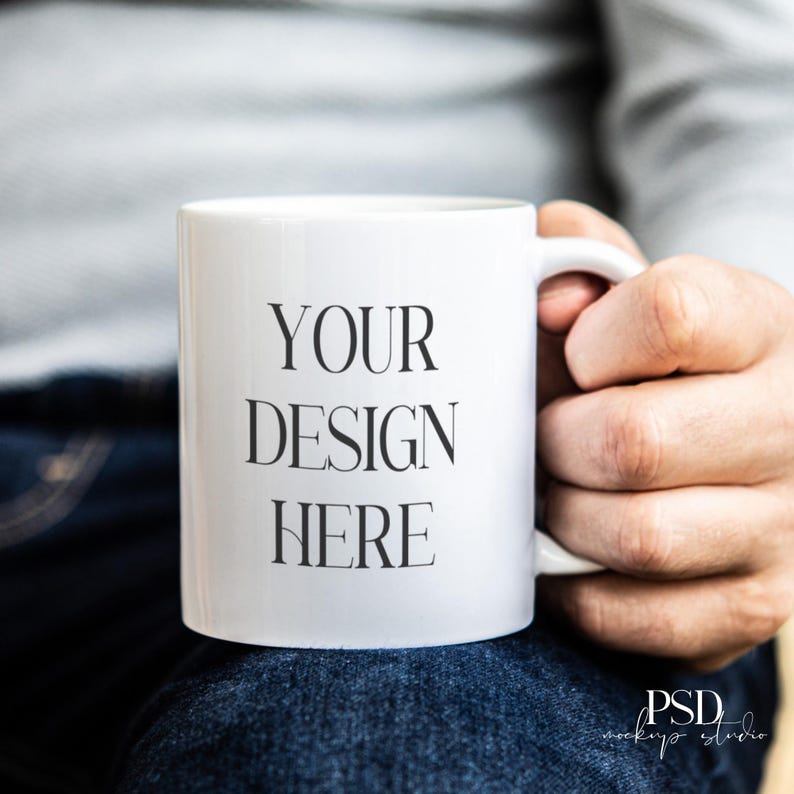 May include: A white ceramic mug with the text "YOUR DESIGN HERE" in black. The mug is held by a person wearing a gray long-sleeve shirt and blue jeans. The mug is a standard size.