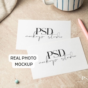 May include: Two white rectangular cards featuring the black text PSD mockup studio, accompanied by a light pink pen, a small blue pin, and a white mug with blue stripes. A white label with the text REAL PHOTO MOCKUP is visible on the left.