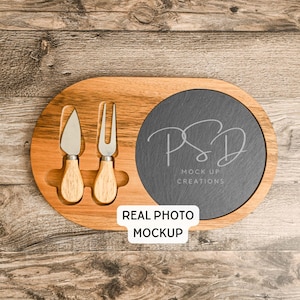May include: Wooden cheese board with a dark gray slate insert and two cheese knives. The board has an oval shape with a cutout for the knives and a round slate area. The text "REAL PHOTO MOCKUP" is visible.