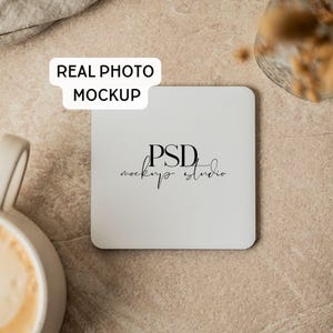 Può includere: Un sottobicchiere quadrato bianco con il testo nero PSD mockup studio al centro. Un'etichetta bianca con il testo nero REAL PHOTO MOCKUP è posizionata sopra il sottobicchiere. L'oggetto è appoggiato su una superficie beige testurizzata.