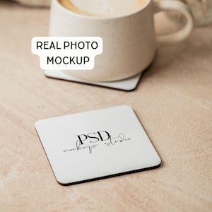 Può includere: Un sottobicchiere quadrato bianco con il testo nero PSD mockup studio al centro. Una tazza in ceramica beige poggia su un altro sottobicchiere dietro quello quadrato. Il testo REAL PHOTO MOCKUP è visualizzato in un riquadro bianco sopra la tazza.