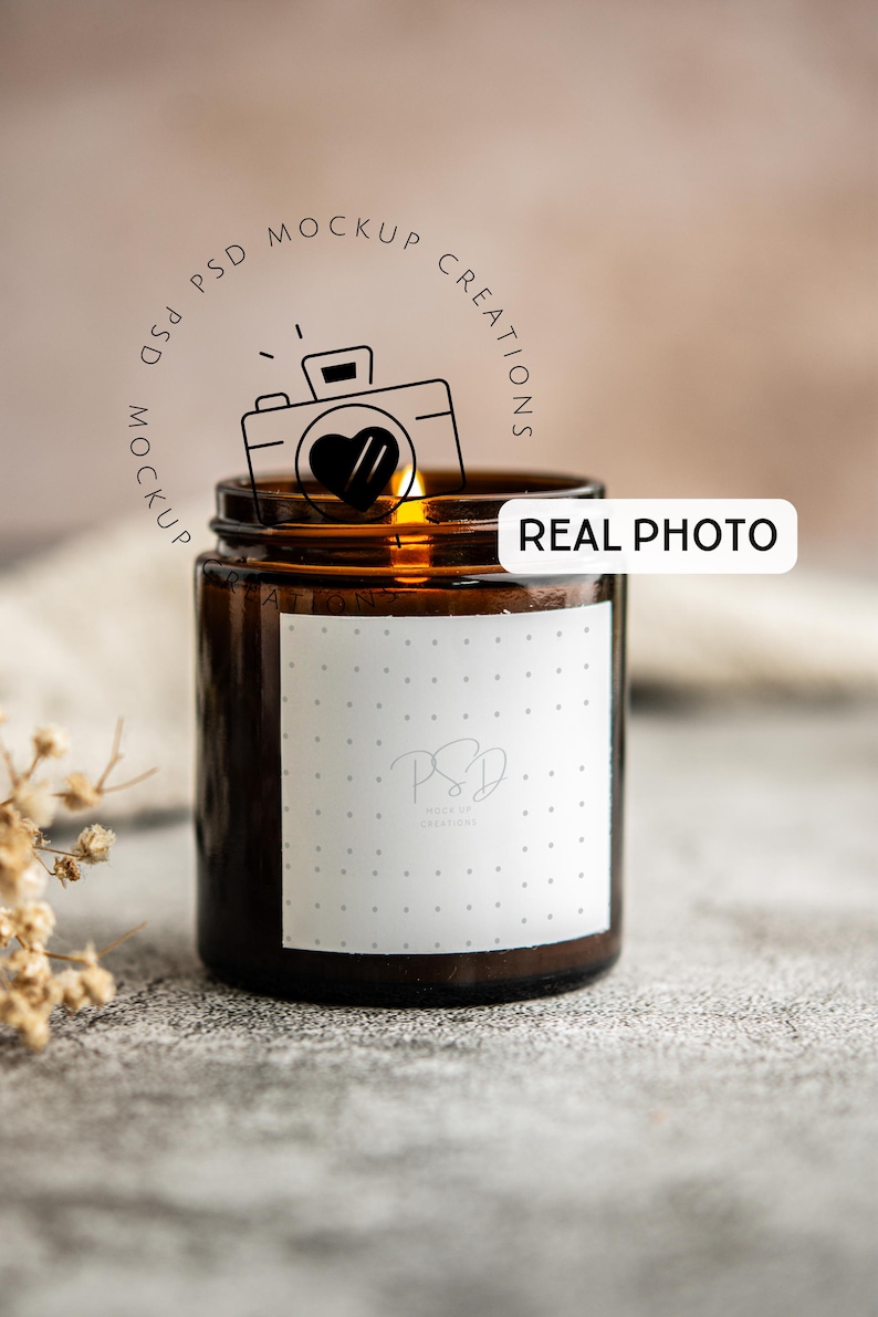 May include: A brown glass jar candle with a white label featuring a logo and dotted pattern. The candle is lit, with a warm glow visible. The words "REAL PHOTO" are on a white rectangle. The image has a neutral background.