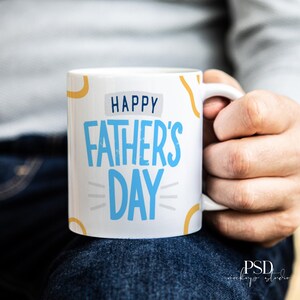 May include: White ceramic mug with the text "HAPPY FATHER'S DAY" in blue and white. The mug has yellow and orange decorative accents. The mug is being held by a person.