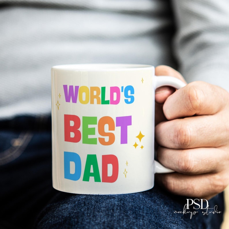 May include: White ceramic mug with the text "WORLD'S BEST DAD" in colorful, bold letters. The mug has a white handle and is held by a person. The design includes star accents.