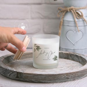 May include: A frosted glass candle with a white label featuring a leafy design and the text "Wedding of Andrea & Oliver 18th November 2019". The candle is on a wooden tray.