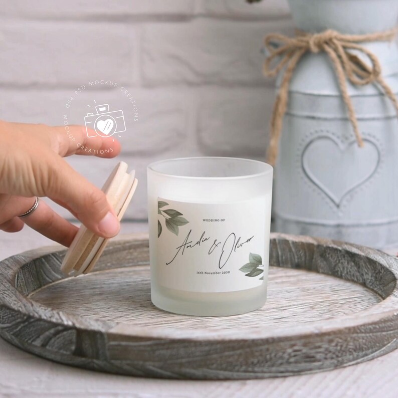 May include: A frosted glass candle with a white label featuring a leafy design and the text "Wedding of Andrea & Oliver 18th November 2019". The candle is on a wooden tray.
