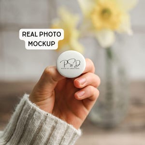 May include: A white round phone grip with the text "PSD MOCK UP CREATIONS" in black script. The phone grip is held in a hand, with a blurred background of flowers and the text "REAL PHOTO MOCKUP".