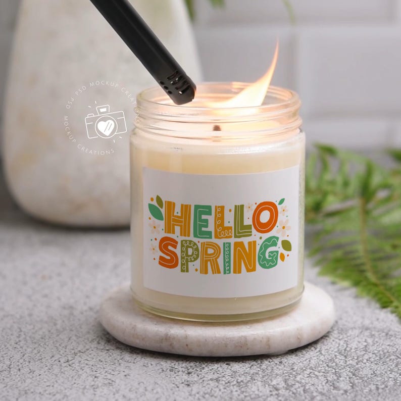 Op de afbeelding: Een witte kaars met een label waarop "Hello Spring" staat in kleurrijke letters. De kaars is aangestoken en de vlam is zichtbaar. De kaars staat op een witte onderzetter.