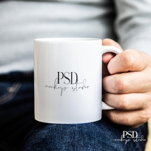 May include: A white ceramic mug with a curved handle. The mug has the text "PSD mockup studio" printed on the front in a black, elegant font. The mug is held by a person.