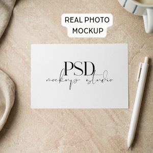 May include: A rectangular white card featuring the black text PSD mockup studio in the center. A white pen is positioned to the right of the card. The card is placed on a textured beige surface next to a piece of beige fabric and a white mug.