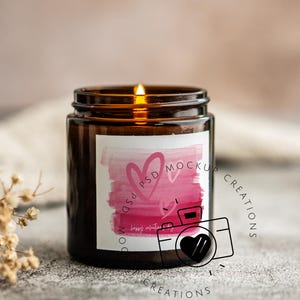 May include: A lit brown glass candle jar with a white label featuring pink watercolor hearts and the text "Happy Valentine's Day". The candle is on a gray surface.