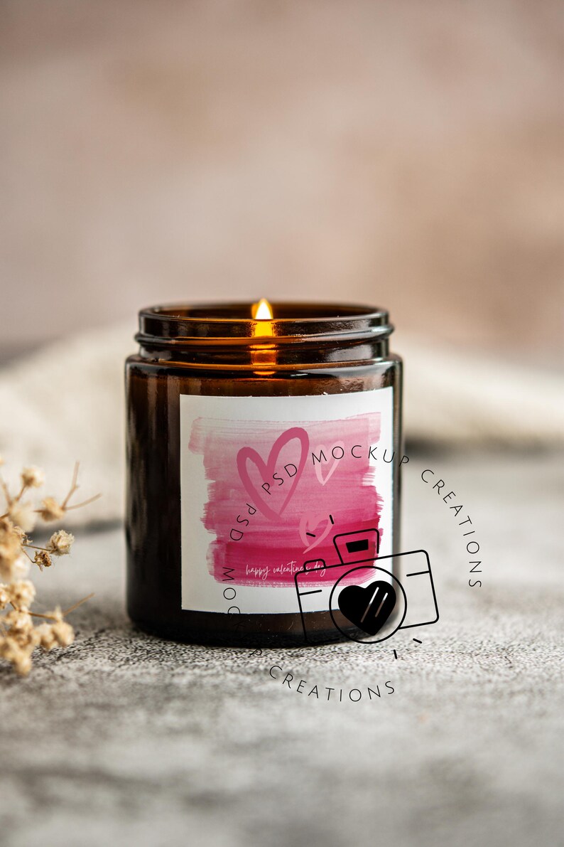 May include: A lit brown glass candle jar with a white label featuring pink watercolor hearts and the text "Happy Valentine's Day". The candle is on a gray surface.