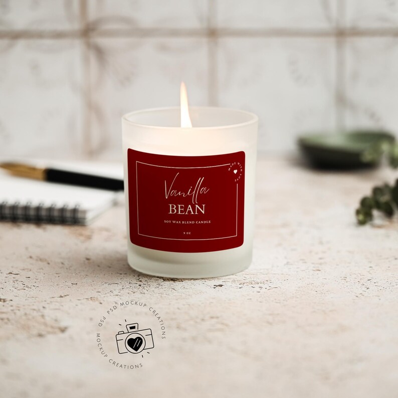 May include: A lit vanilla bean scented soy wax blend candle in a frosted glass jar. The candle has a red label with gold text that reads "Vanilla Bean". The candle is 3 oz. A notebook and pen are in the background.