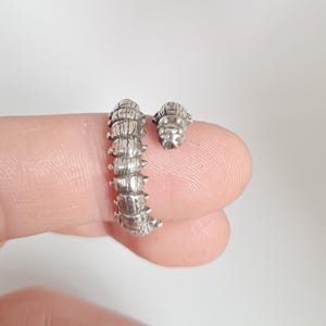 May include: A silver-toned ring shaped like a segmented caterpillar with a textured, ridged body and a small head piece. The design features an open-ended, wrap-around style that curves to follow the shape of a finger, showcasing metallic details.