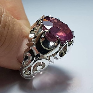 Peut inclure: Une bague en argent ornée d'une pierre précieuse ovale, facettée, rouge-violet. La bague présente un design ajouré orné de motifs tourbillonnants. La pierre précieuse est le point central, captant la lumière et mettant en valeur sa couleur et sa taille.