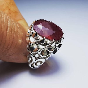 Peut inclure: Une bague en argent ornée d'une pierre précieuse ovale, facettée, de couleur rouge foncé. Le sertissage de la bague présente un motif floral détaillé et texturé. La bague est présentée sur un fond clair.