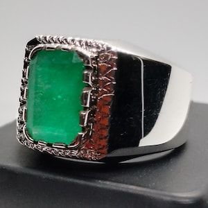 Peut inclure: Une bague en argent ornée d'une grande pierre verte émeraude rectangulaire. La pierre est sertie dans un cadre décoratif et texturé avec des accents brun-rouge. La bague a une surface polie et réfléchissante.