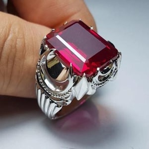 Peut inclure: Une bague en argent ornée d'une grande pierre précieuse rectangulaire rouge foncé. La bague présente un design orné avec des détails complexes autour de la pierre et sur le bandeau. La pierre est facettée, reflétant la lumière.