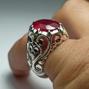 May include: A silver ring with an oval, deep red gemstone. The ring features an ornate, filigree design around the stone and band. The gemstone is the focal point, catching the light and adding a touch of elegance to the jewelry.