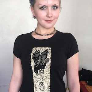 K&ouml;nnte beinhalten: Schwarzes T-Shirt mit einem grafischen Aufdruck eines Rabens, der eine Flasche mit zwei Herzen h&auml;lt. Die Flasche steht in Flammen und der Text "Liv Rainney-Smith" befindet sich am unteren Rand des Designs.