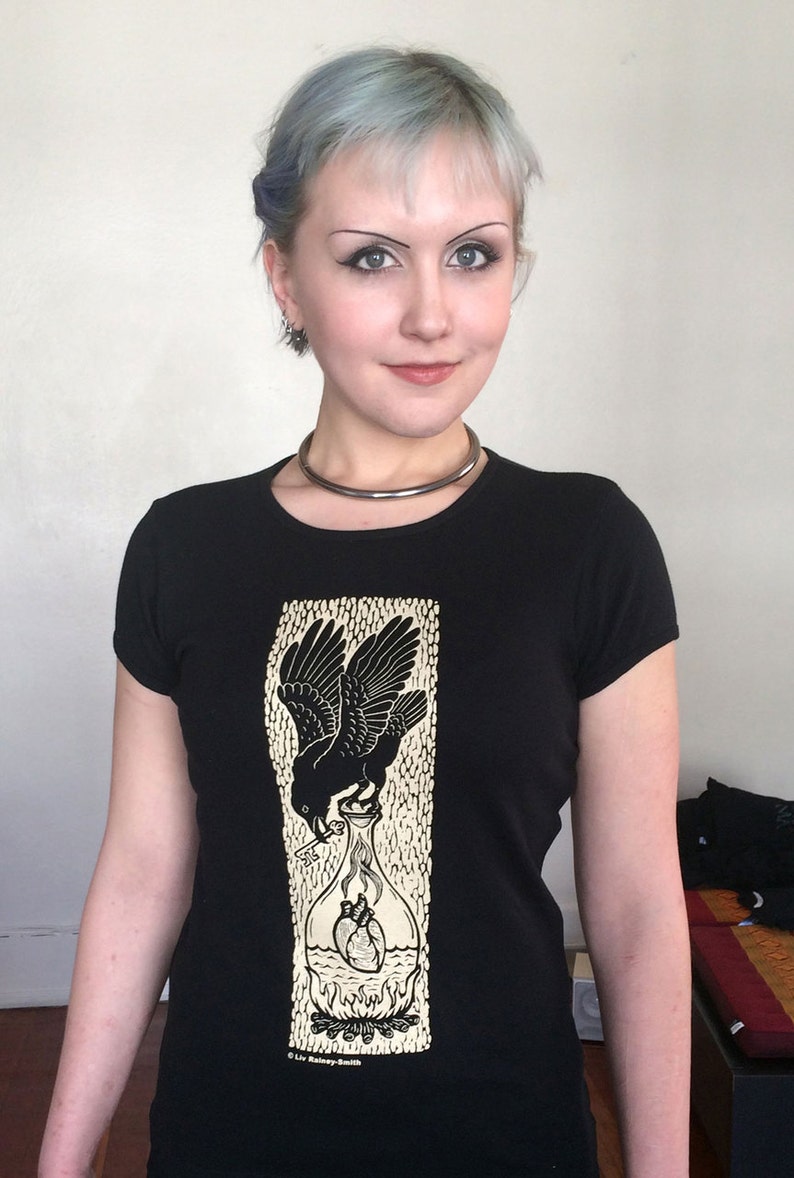 K&ouml;nnte beinhalten: Schwarzes T-Shirt mit einem grafischen Aufdruck eines Rabens, der eine Flasche mit zwei Herzen h&auml;lt. Die Flasche steht in Flammen und der Text "Liv Rainney-Smith" befindet sich am unteren Rand des Designs.