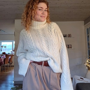 May include: A white cable knit jumper with a turtleneck and long sleeves. The jumper has a loose fit and is worn with beige trousers and a brown belt.