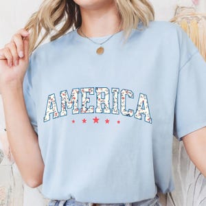 May include: Pale blue t-shirt featuring the word "AMERICA" in a floral pattern. The word is above five red stars. The t-shirt has a crew neck and short sleeves. The floral design includes red and white flowers.