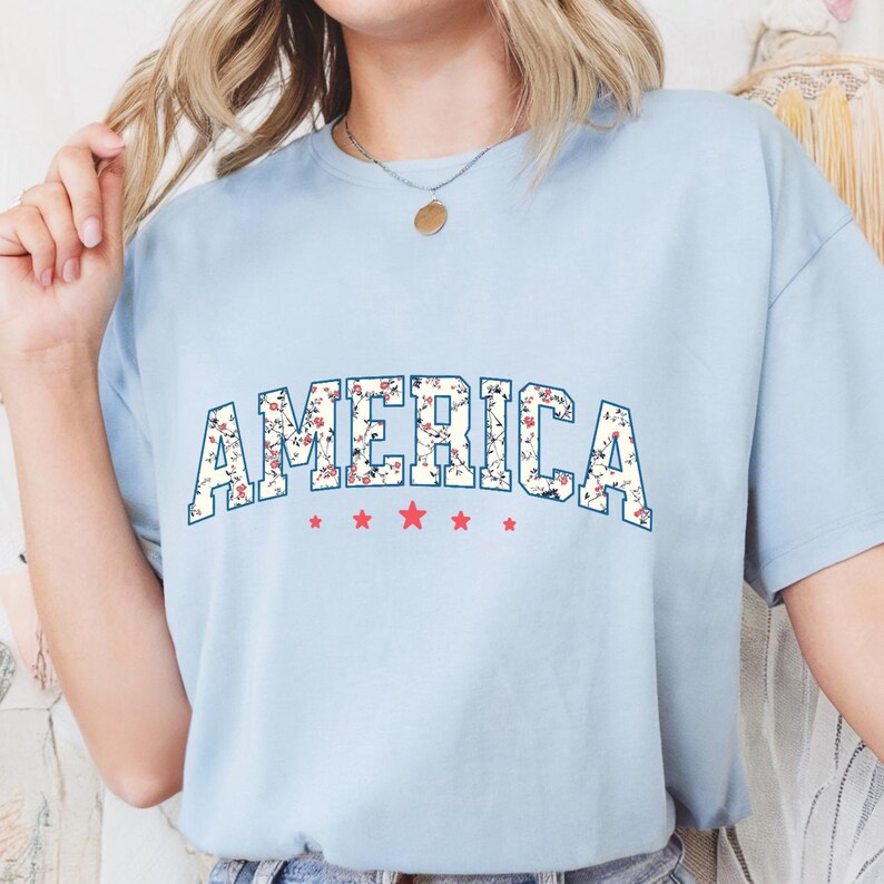 May include: Pale blue t-shirt featuring the word "AMERICA" in a floral pattern. The word is above five red stars. The t-shirt has a crew neck and short sleeves. The floral design includes red and white flowers.