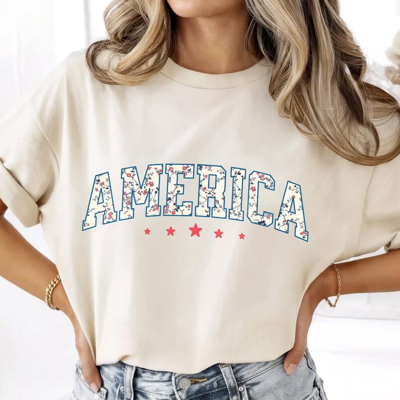 May include: A cream-coloured t-shirt featuring the word "AMERICA" in a blue outline, floral-patterned font. Below the word are five red stars. The shirt is a casual style, suitable for patriotic events.