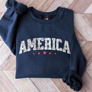 May include: A navy blue sweatshirt featuring the word "AMERICA" in a floral pattern. Beneath the word are five red stars. The garment is displayed on a wooden surface.