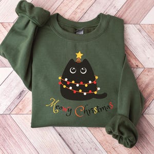 May include: Green sweatshirt with a black cat dressed as a Christmas tree. The cat has a star on its head and is wearing a string of Christmas lights. The text "Meowy Christmas" is printed below the cat.