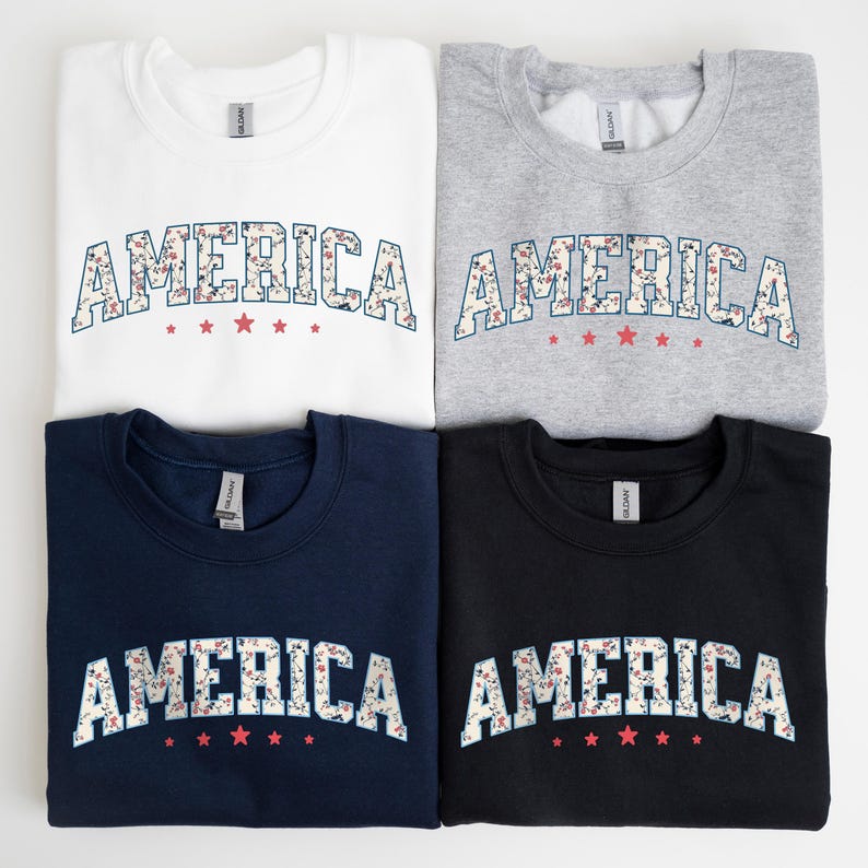 May include: Four crew neck sweatshirts in white, grey, navy, and black. Each sweatshirt displays the word "AMERICA" in a floral design with red star accents. The sweatshirts are folded and displayed on a white surface.