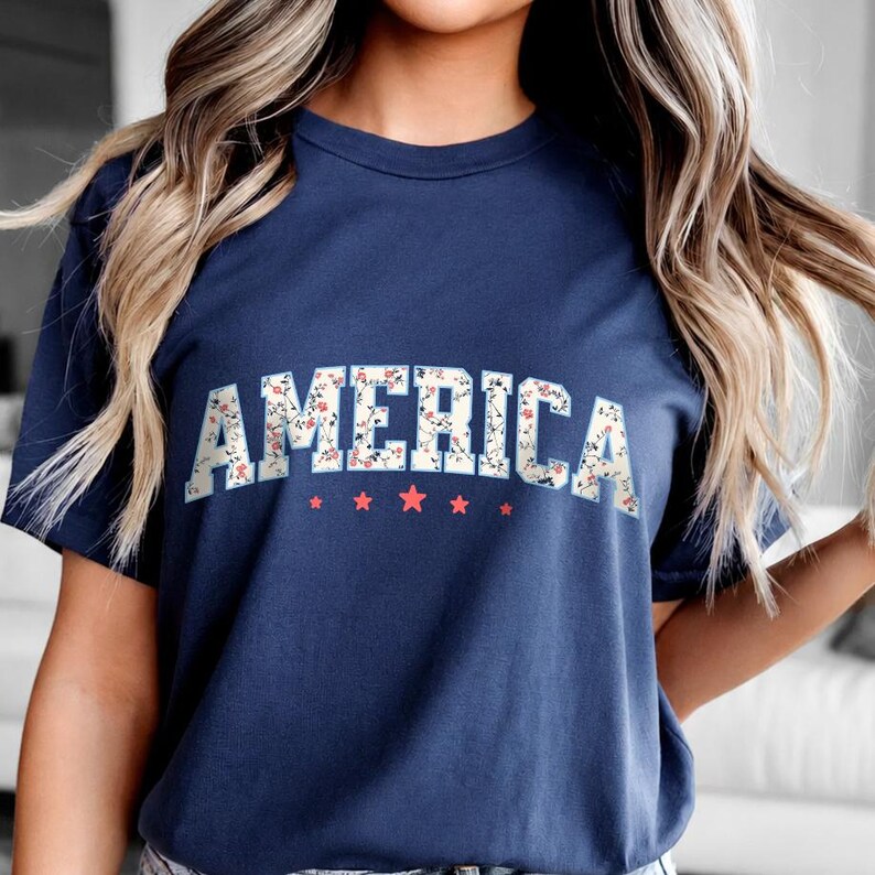 May include: A navy blue t-shirt featuring the word "AMERICA" in a floral design. Beneath the word are five red stars. The shirt has a crew neck.