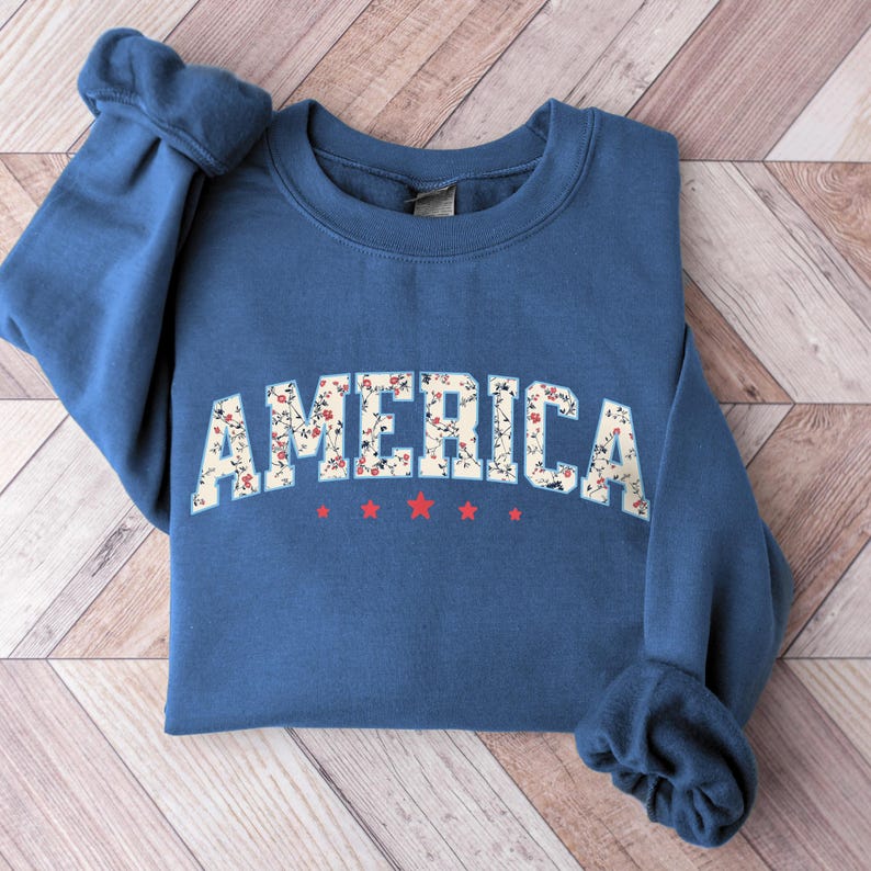 May include: A blue sweatshirt featuring the word "AMERICA" in a floral pattern. Beneath the word are three red stars. The sweatshirt has a crew neck and long sleeves.