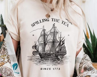 May include: Cream-colored t-shirt featuring a detailed black and white illustration of a tall ship on the sea. The shirt has the text "Spilling the Tea Since 1773" printed above and below the ship graphic. The shirt is short-sleeved.