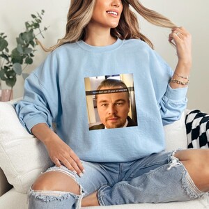 May include: A light blue sweatshirt with a graphic of Leonardo DiCaprio's face and the text "How don't I turn 15 if you're so very old".