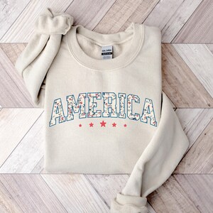 May include: Cream-coloured sweatshirt featuring the word "AMERICA" in a floral design outlined in blue. Below the word are three red stars. The sweatshirt is folded on a wooden surface.