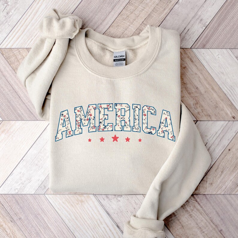 May include: Cream-coloured sweatshirt featuring the word "AMERICA" in a floral design outlined in blue. Below the word are three red stars. The sweatshirt is folded on a wooden surface.